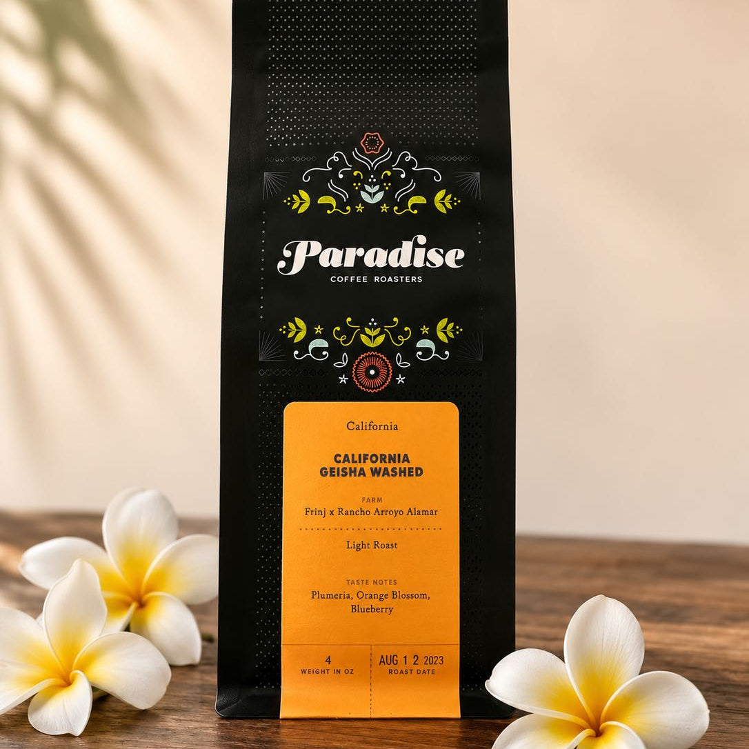A bag of Paradise Coffee Roasters California geisha washed coffee stands upright on a wooden surface, flanked by white plumeria flowers against a warm beige background with soft leaf shadows.