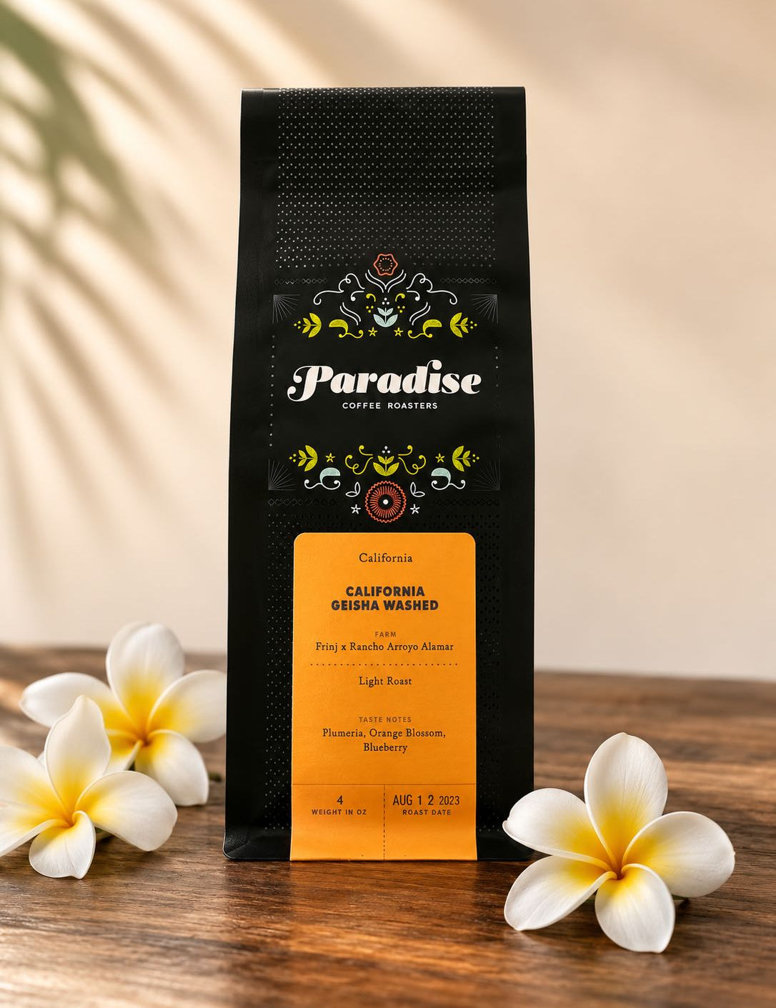 A bag of Paradise Coffee Roasters California geisha washed coffee stands upright on a wooden surface, flanked by white plumeria flowers against a warm beige background with soft leaf shadows.