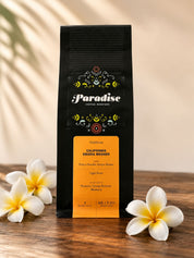 A bag of Paradise Coffee Roasters California geisha washed coffee stands upright on a wooden surface, flanked by white plumeria flowers against a warm beige background with soft leaf shadows.