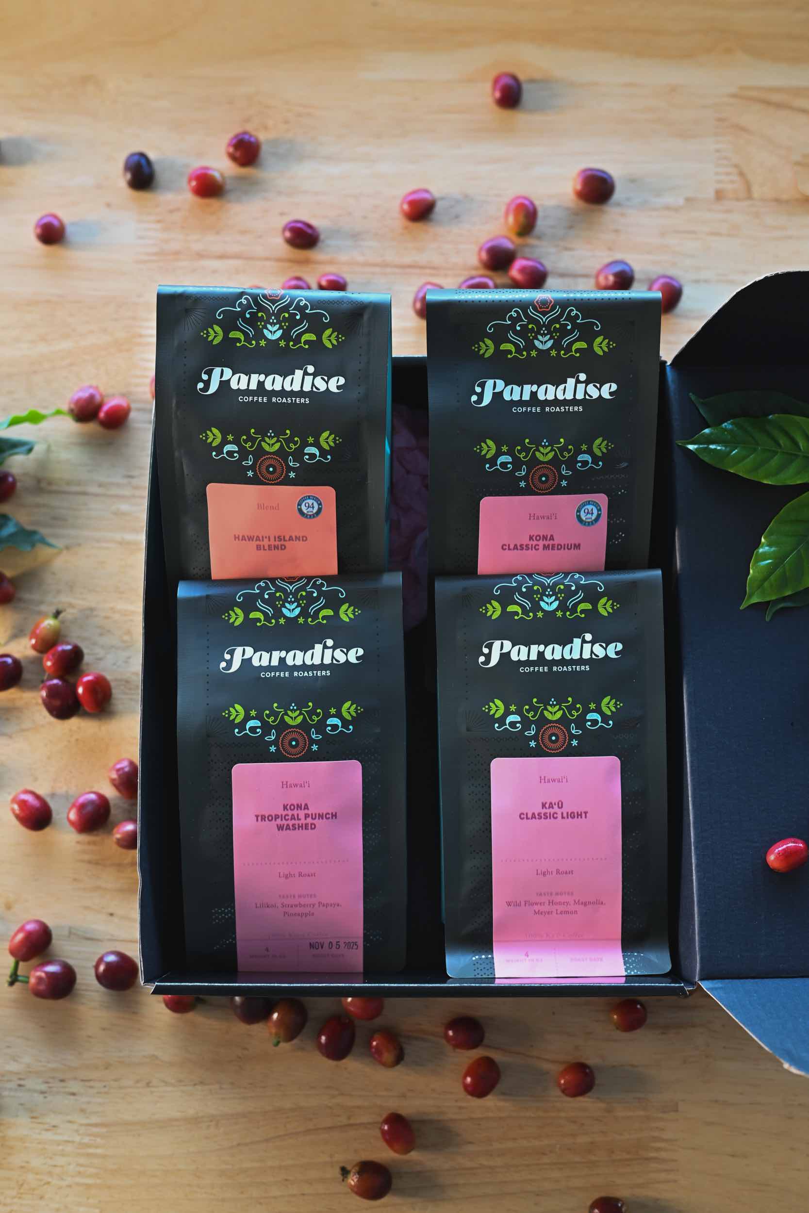 Four Paradise Roasters coffee bags in a box with scattered coffee beans on a wooden surface.